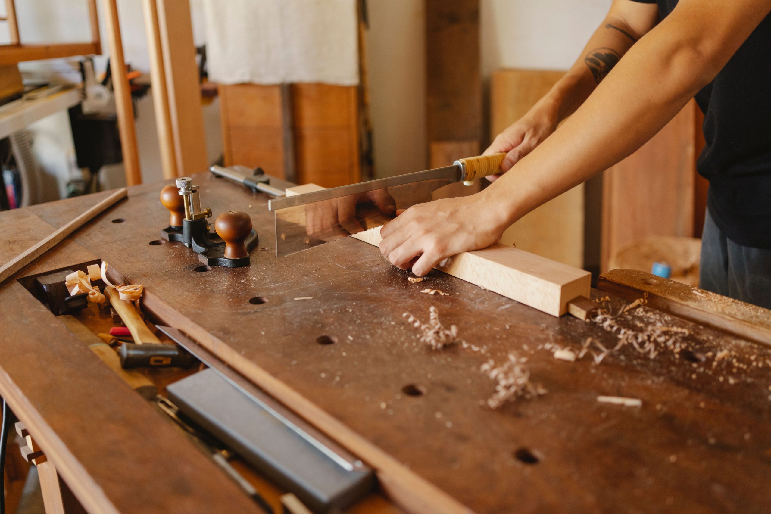Crop anonymous craftsman with tattoo sawing wooden board with hacksaw at workplace in carpentry workshop